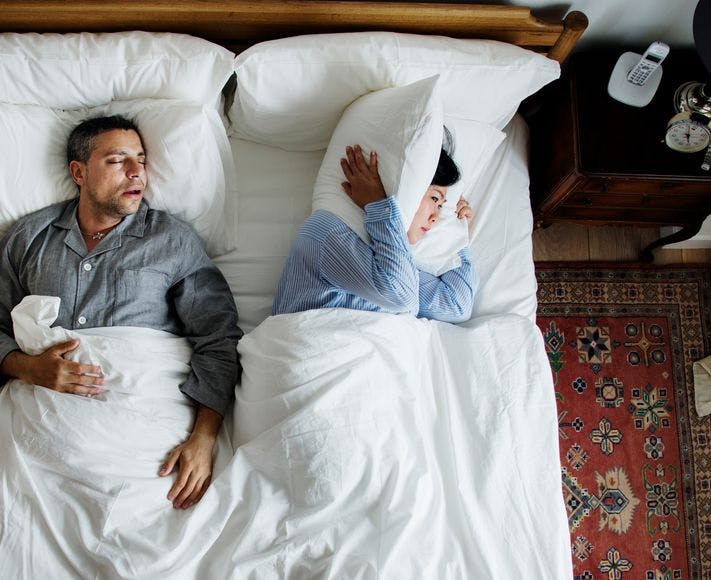 An man sleeping and snoring in bed with a woman lying sideways, covering her ears with a pillow.