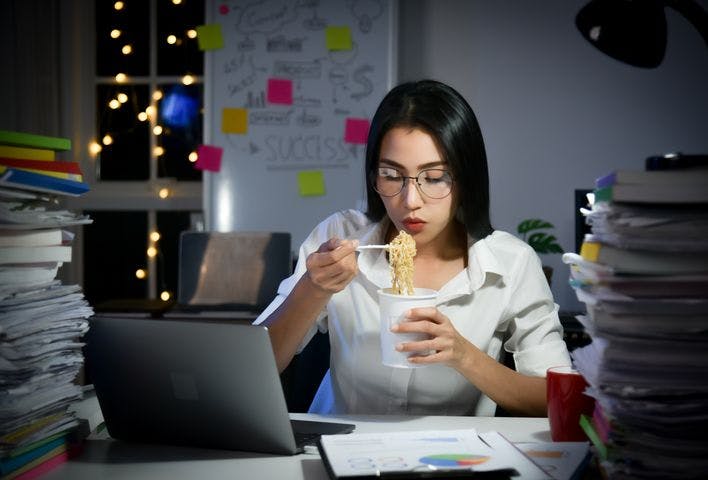 A female employee eating instant cup noodles at her work desk, surrounded by stacks of papers.