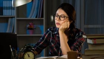 A woman looking dazed while staring at a laptop; a clock showing 5 minutes to midnight on her desk.