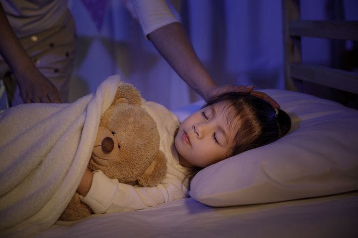 A child asleep in bed while holding a teddy bear as a woman caresses her head with her left hand