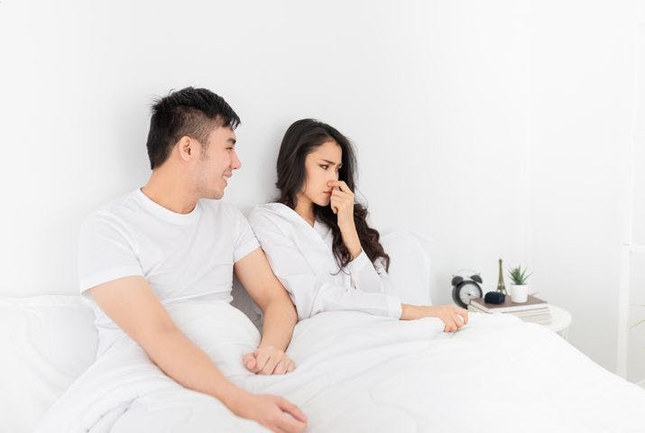 Man looking at a woman smilingly as she looks to the side while closing her nose as they sit up in bed