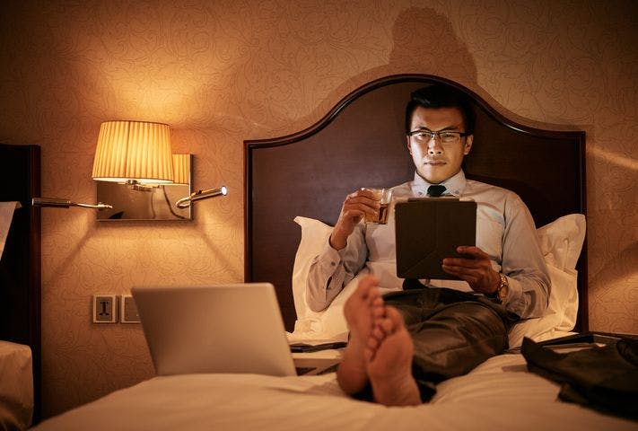 A man in business attire sitting up on a bed while holding a tablet in one hand and a glass of rum in another.