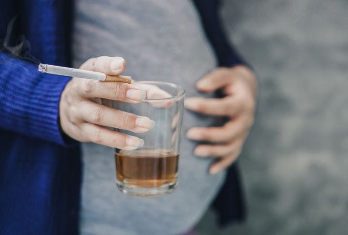 Pregnant woman holding a cigarette and cup of beverage in her right hand while placing her left hand on her abdomen