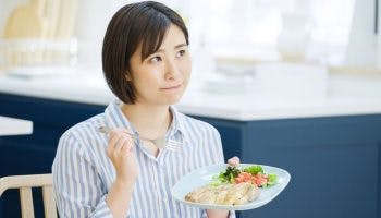 A woman thinking while holding a plate of food in one hand and a fork in the other hand.  