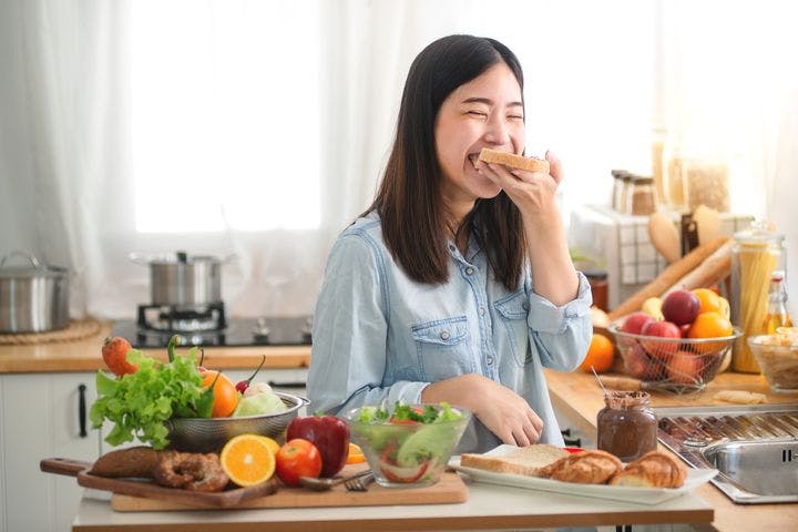 A woman biting into a slice of bread while being surrounded by fruits and vegetables in her kitchen.