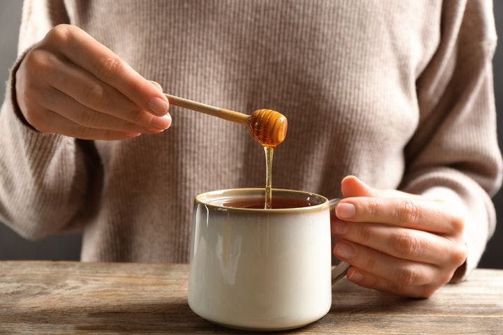 A partial view of a woman’s hands inserting honey into a drinking mug.