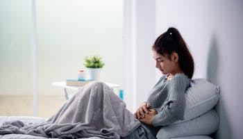 A woman sitting up on her bed while holding a part of her stomach in pain.