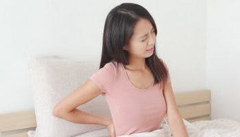A woman sitting on her bed holding her lower back in pain.