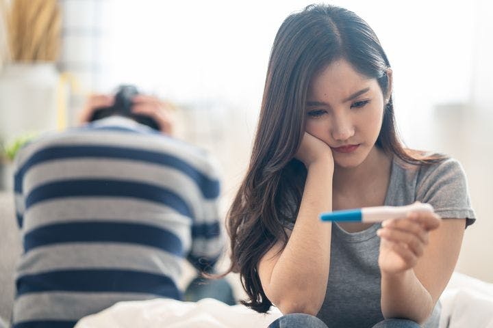 A woman looking disappointingly at a pregnancy test with the back of a frustrated man in the background.