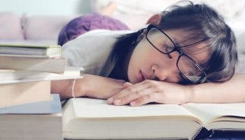 Woman with glasses asleep with her face and hands resting on a book