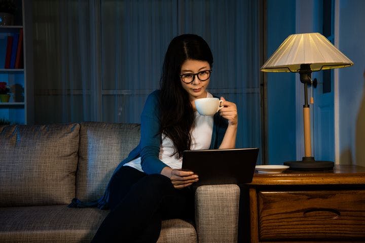 A woman holding a cup of tea in one hand while watching something on her gadget.