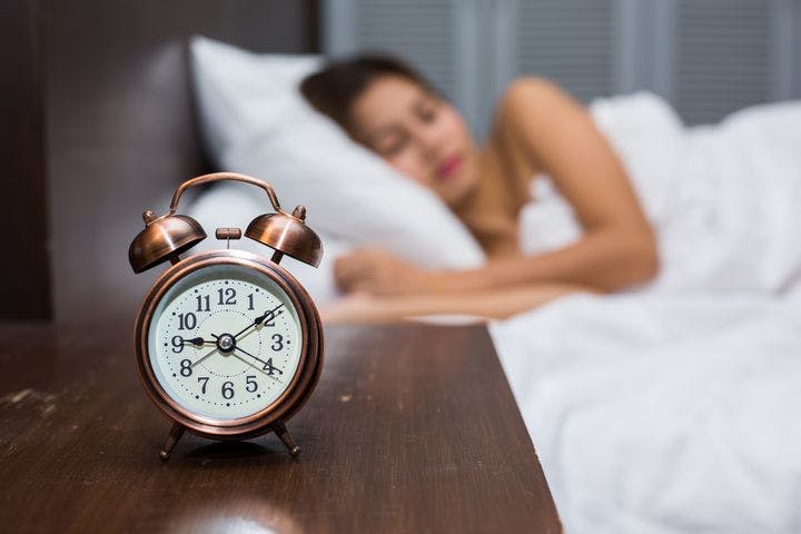 A table clock with a blurred image of a woman sleeping on her bed in the background.