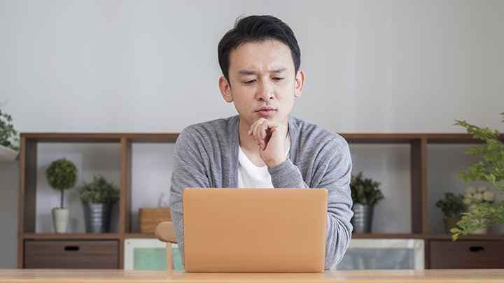 An Asian man looking confused, staring at his laptop in the living room