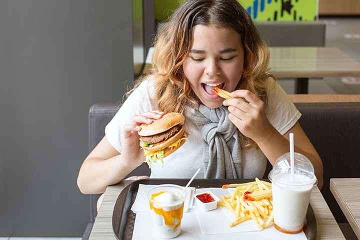 Girl eating French fries dipped in chilli sauce with her left hand and holding a burger in her right