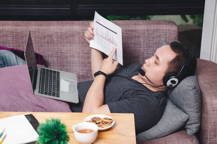 An Asian man lying down on a couch while doing online presentation using his laptop and headphone