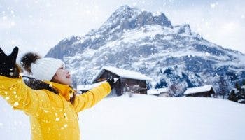 Woman wearing a yellow winter coat and white winter hat enjoys snow with a mountain in the background in a cold-weather country