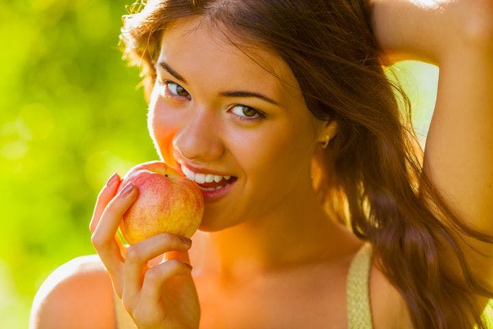 Woman holding an apple close to her lips as she smiles while holding the back of her head with her left hand 
