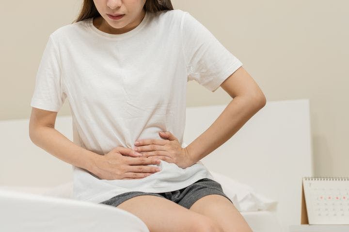 A partial view of a woman sitting on her bed and holding her stomach in pain.