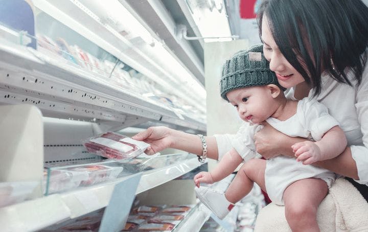 A mother carrying her infant while picking out frozen ingredients in a supermarket.