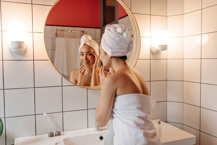 A woman standing looking at herself in a bathroom mirror wearing a towel and looking satisfied while touching her face