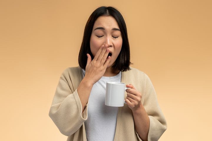 Woman in a dressing gown yawning with eyes closed while covering her mouth her right hand and holding a mug in her left