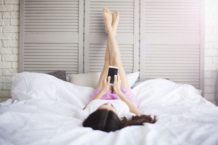 A woman lying in bed with her legs raised straight up and messaging on her phone.