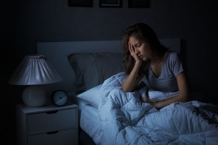 Woman sitting up in bed with closed eyes and her right hand covering her right eye 