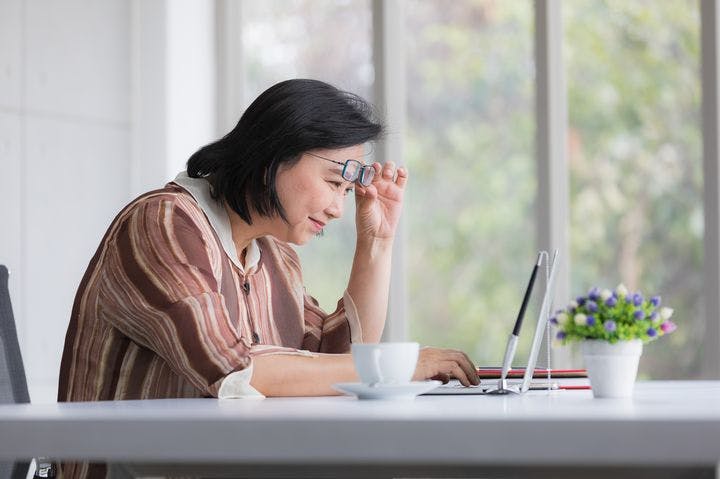 Woman lifting her eyeglasses to look at her laptop while sitting at a table
