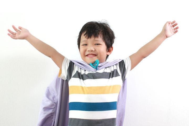 A young boy smiling and throwing his hands up while wearing a sheet as a superhero cape.
