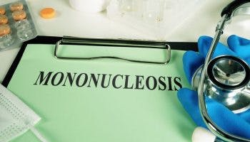 The word “mononucleosis” is printed on a green pad placed on the table next to medical gloves, stethoscope, surgical mask, pills, and test tube rack.