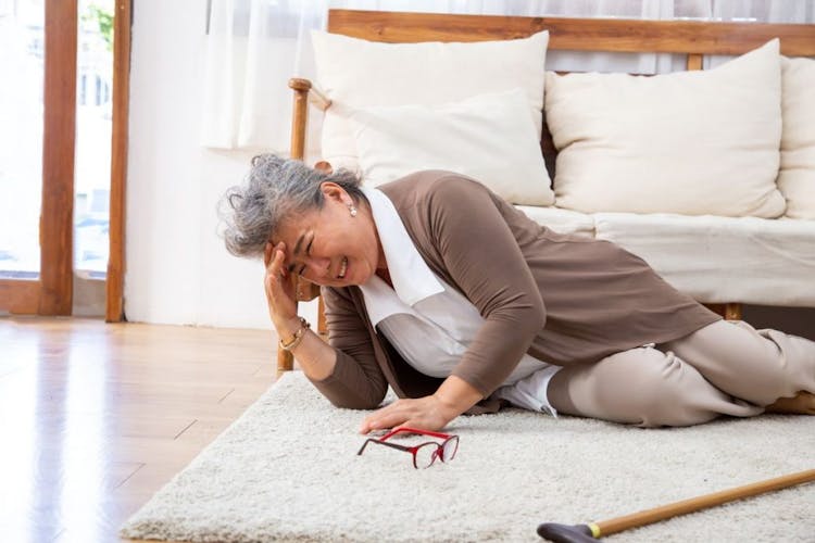 Woman falling down on a rug while holding her forehead as a pair of eyeglasses and wooden cane lie next to her