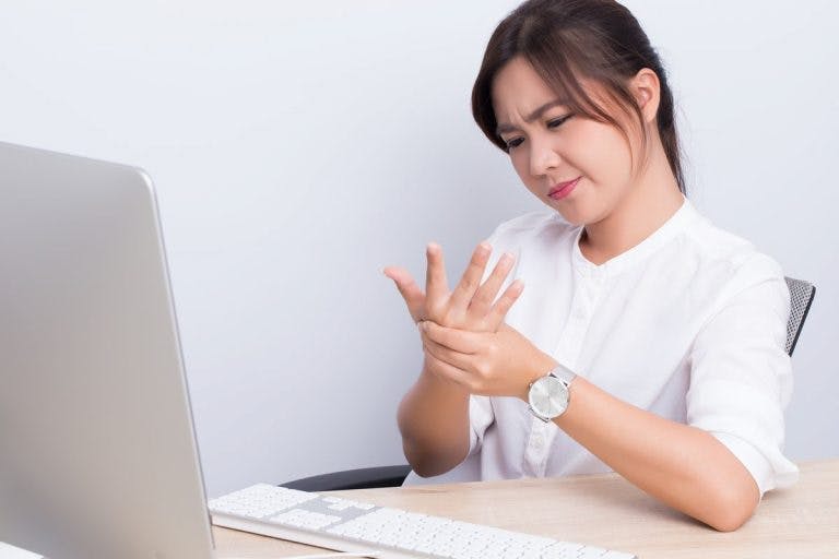 Woman rubbing her right hand with her left as she sits in front of a keyboard and desktop computer.