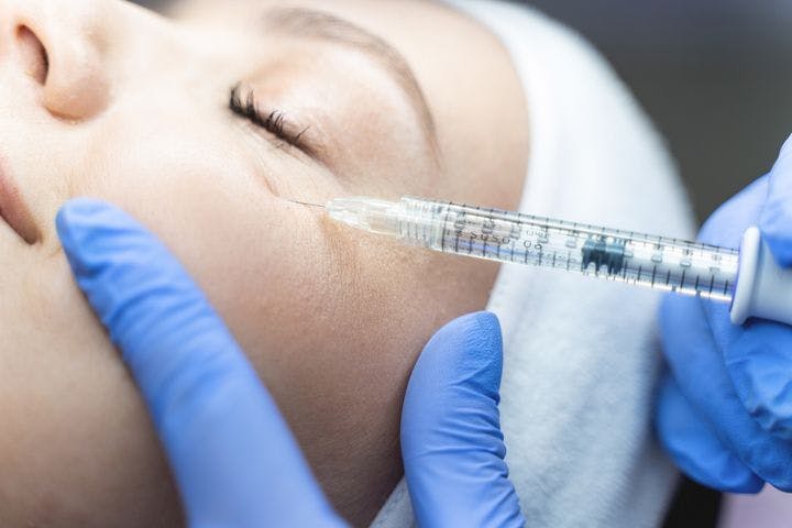 Closeup of hands wearing medical grade gloves injecting fillers into a woman’s tear trough area