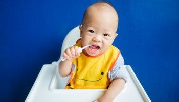 A baby sitting in a baby chair while holding a toothbrush and placing it in his mouth