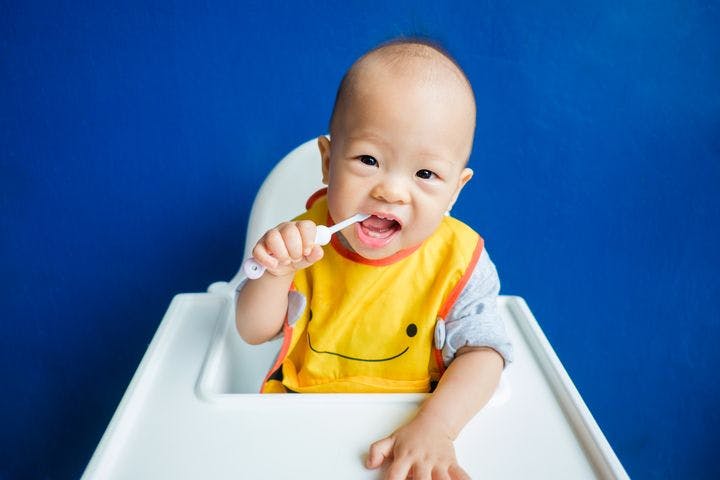 A baby sitting in a baby chair while holding a toothbrush and placing it in his mouth