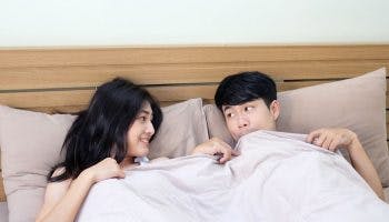 Woman smiling at a man who’s biting a blanket while looking at her as they sit up in bed 