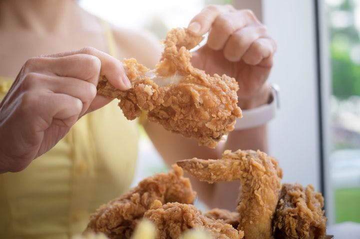 A partial view of a woman tearing apart a piece of fried chicken with her hands.