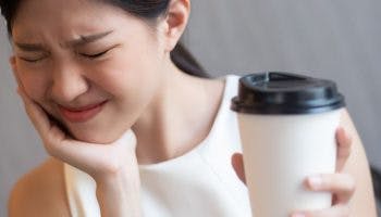 Woman wincing in pain while supporting her chin with her right hand and holding a cup of coffee in her left