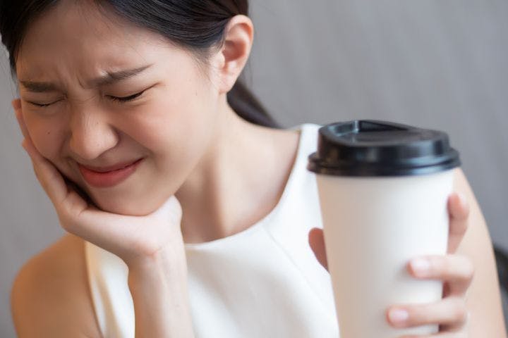 Woman wincing in pain while supporting her chin with her right hand and holding a cup of coffee in her left