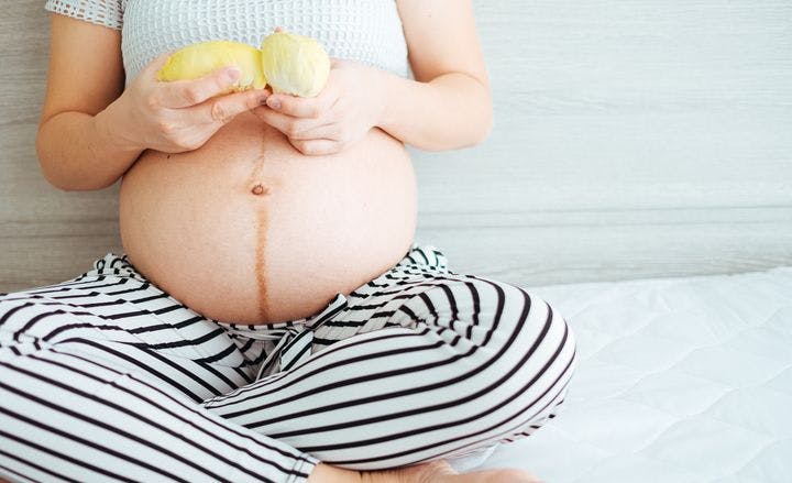 Close-up of a pregnant women with an exposed belly sitting cross-legged while holding a durian pulp in each hand