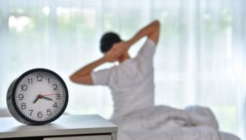 Man waking up by stretching his body facing the window, a clock shows it’s 7am
