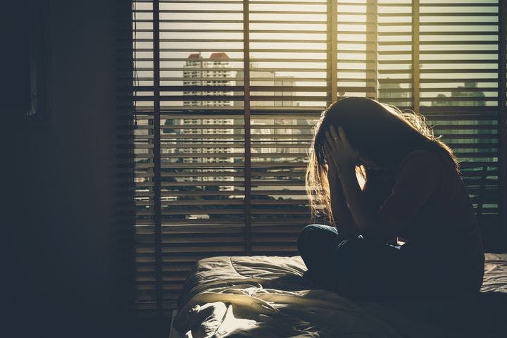 Woman sitting on a bed with her hands in her head as sunlight comes through window blinds