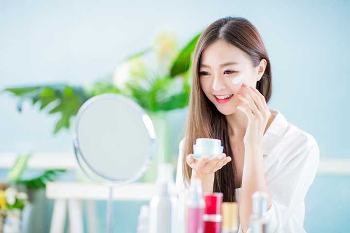 Woman applies facial cream on her face while holding a mirror in one hand.