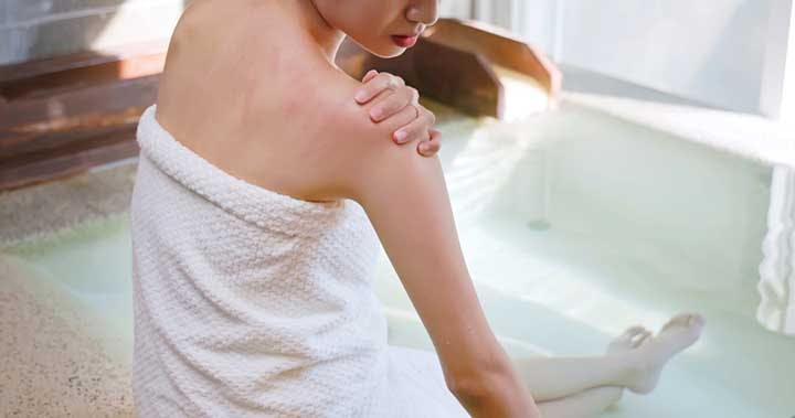 Woman turning to look at streaks of redness on her right shoulder while sitting on the edge of a bathtub