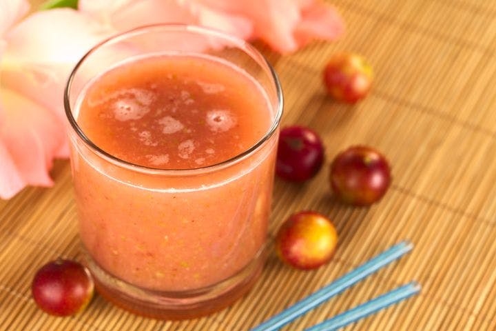 A glass of camu-camu or Myrciaria dubia juice, surrounded by a few loose camu-camu fruits and two blue straws on a bamboo placemat