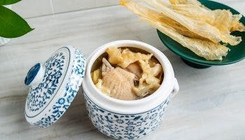 Fish maw soup cooked with chicken in a floral ceramic container alongside a green bowl with dried fish maw.