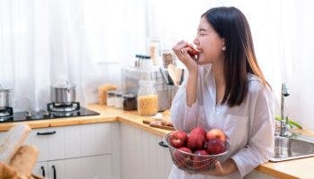 A woman stands in the kitchen; one hand holds a bowl of apples while another hand is holding an apple near her mouth.