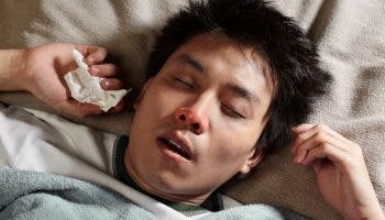 Man sleeps in bed while holding a tissue in one hand, with his mouth slightly agape and the tip of his nose a bit reddish.