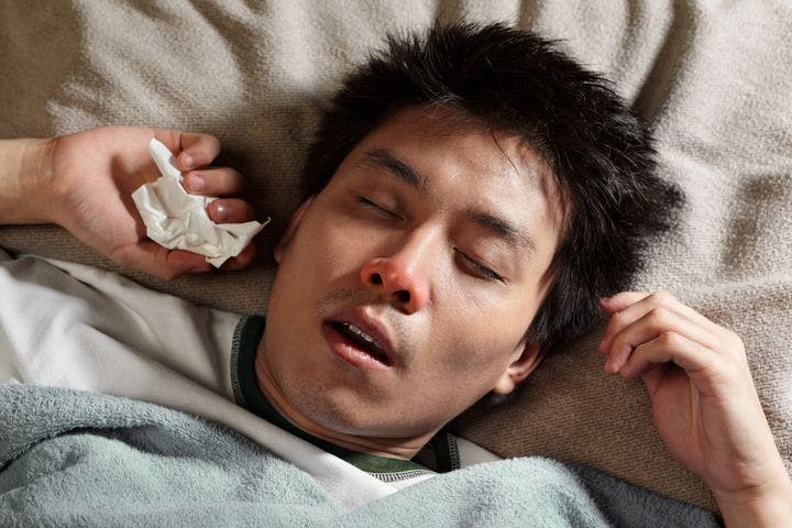 Man sleeps in bed while holding a tissue in one hand, with his mouth slightly agape and the tip of his nose a bit reddish.