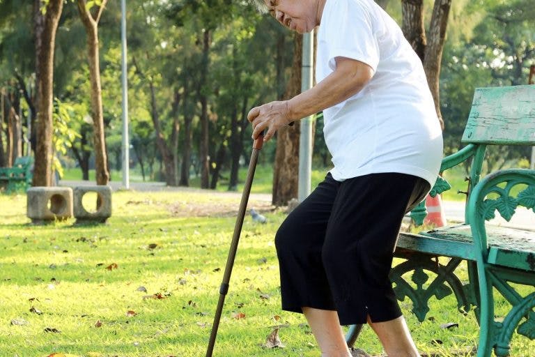 Woman grimacing in pain as she attempts to sit on a wooden park bench while holding a cane in her left hand